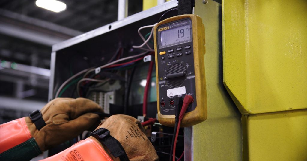 Electrician in insulating gloves testing an equipment enclosure with a multimeter